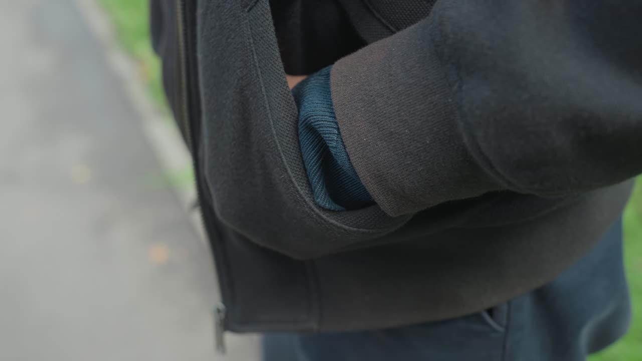 hand exiting jacket pocket near curb, closeup of knit cuff and zippered fleece, subtle movement as pedestrian adjusts clothing, shallow depth emphasizing texture and motion, neutral urban park setting