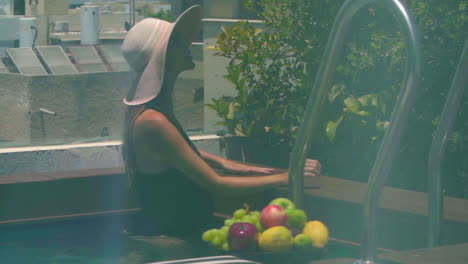 Glass-reflection-shot-of-a-woman-in-a-black-swimsuit-and-straw-hat-walking-by-a-private-pool-with-a-beautiful-view-of-the-city