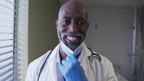 portrait of smiling african american male doctor wearing face mask standing in hospital room