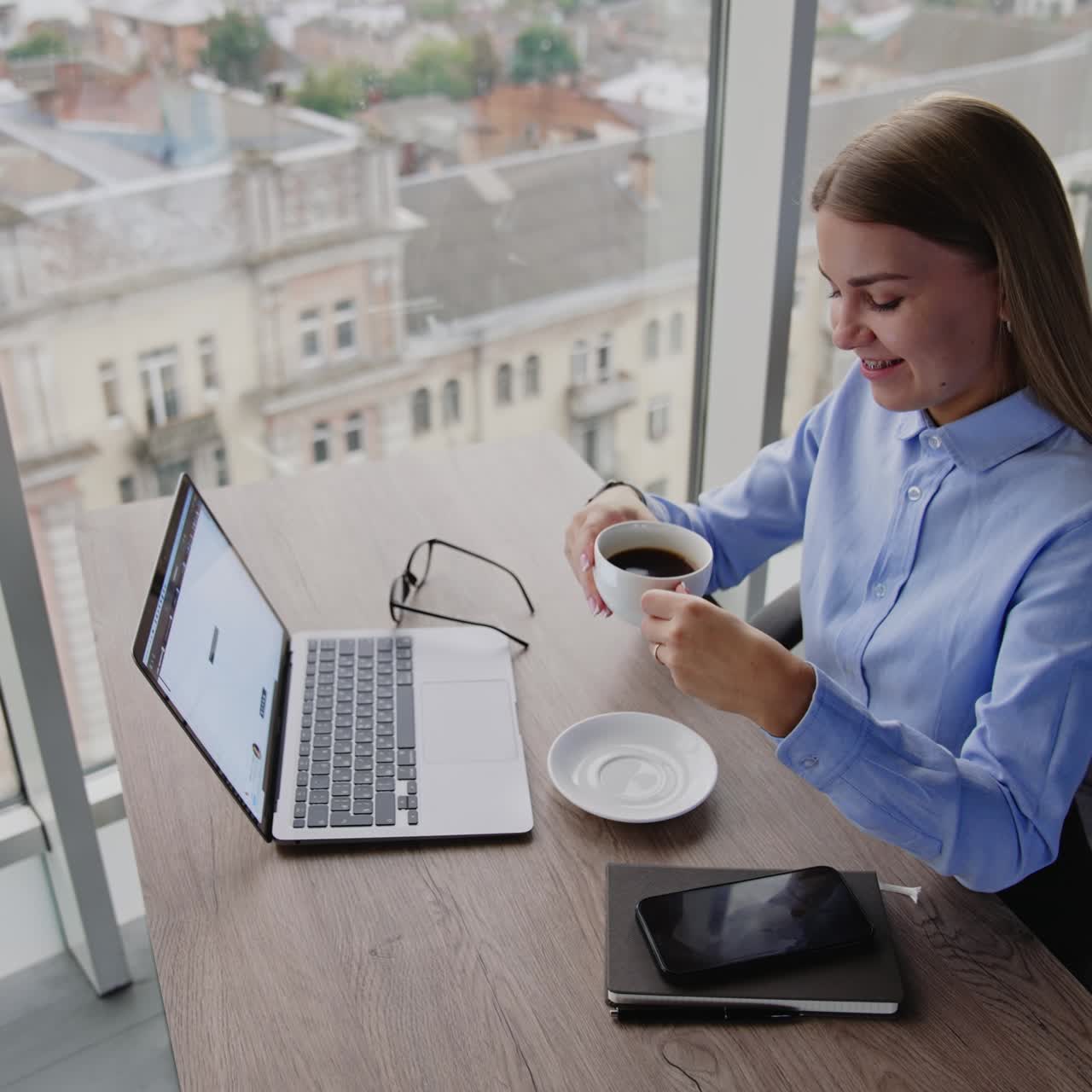 Giovane donna felice e resiliente beve un caffè. La donna guarda il suo portatile e poi si gode la vista dalla finestra. Prospettiva da un’angolazione elevata