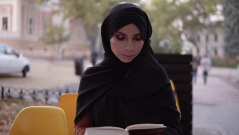 Girl-In-Black-Hijab-Reading-Book-While-Sitting-In-Outdoors-Cafe,-Preparing-For-Exam,-Education-Among-Muslim-Women