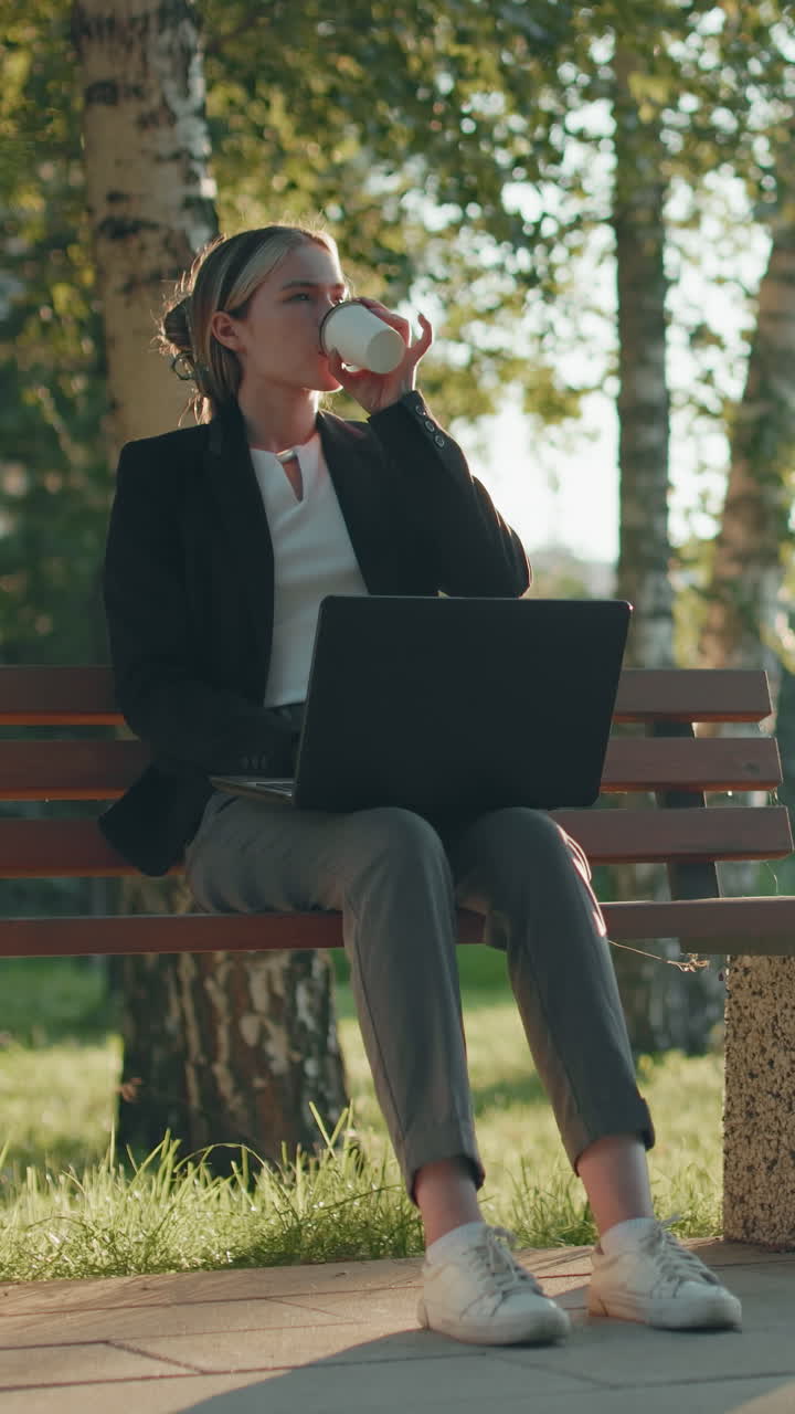Commercialista da remoto in blazer intento a bilanciare il conto usando un laptop mentre è seduto su una panchina di legno in un parco all'aperto con una tazza di caffè di lato e uno sfondo urbano con edifici