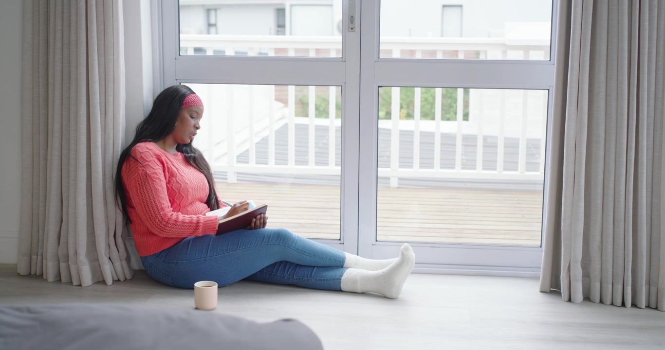 Donna afroamericana che apre un diario color bordeaux, scrivendo sul diario a terra accanto alla porta del balcone, sorseggiando da una tazza.