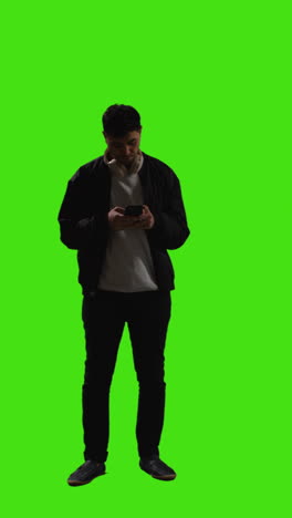 Vertical-Video-Full-Length-Shot-Of-Man-Wearing-Wireless-Headphones-Text-Messaging-On-Mobile-Phone-Against-Green-Screen-1