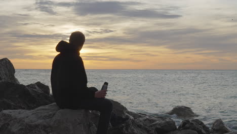 man-drinks-from-cup-near-sea,-closes-thermos,-then-leaves-over-rocks-during-golden-hour