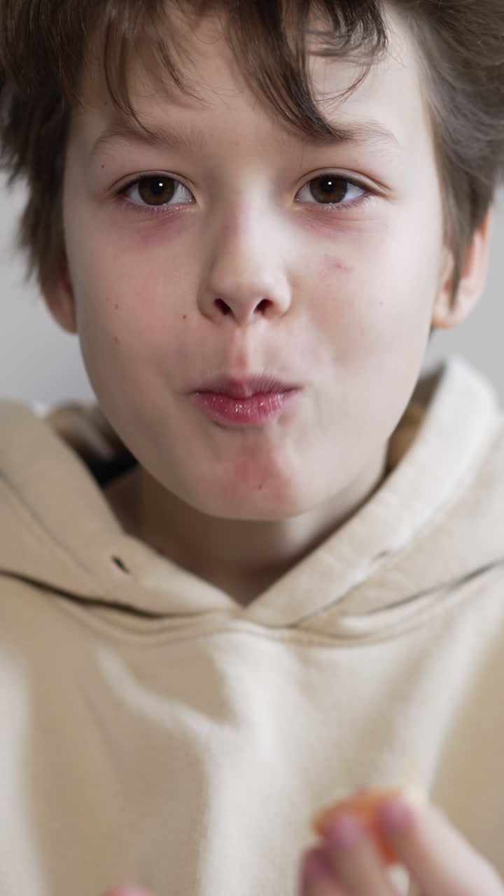 Cute boy with dark disheveled hair eats a juicy tangerine. Kid puts the fruit slices one by one into mouth. Close up. Vertical video