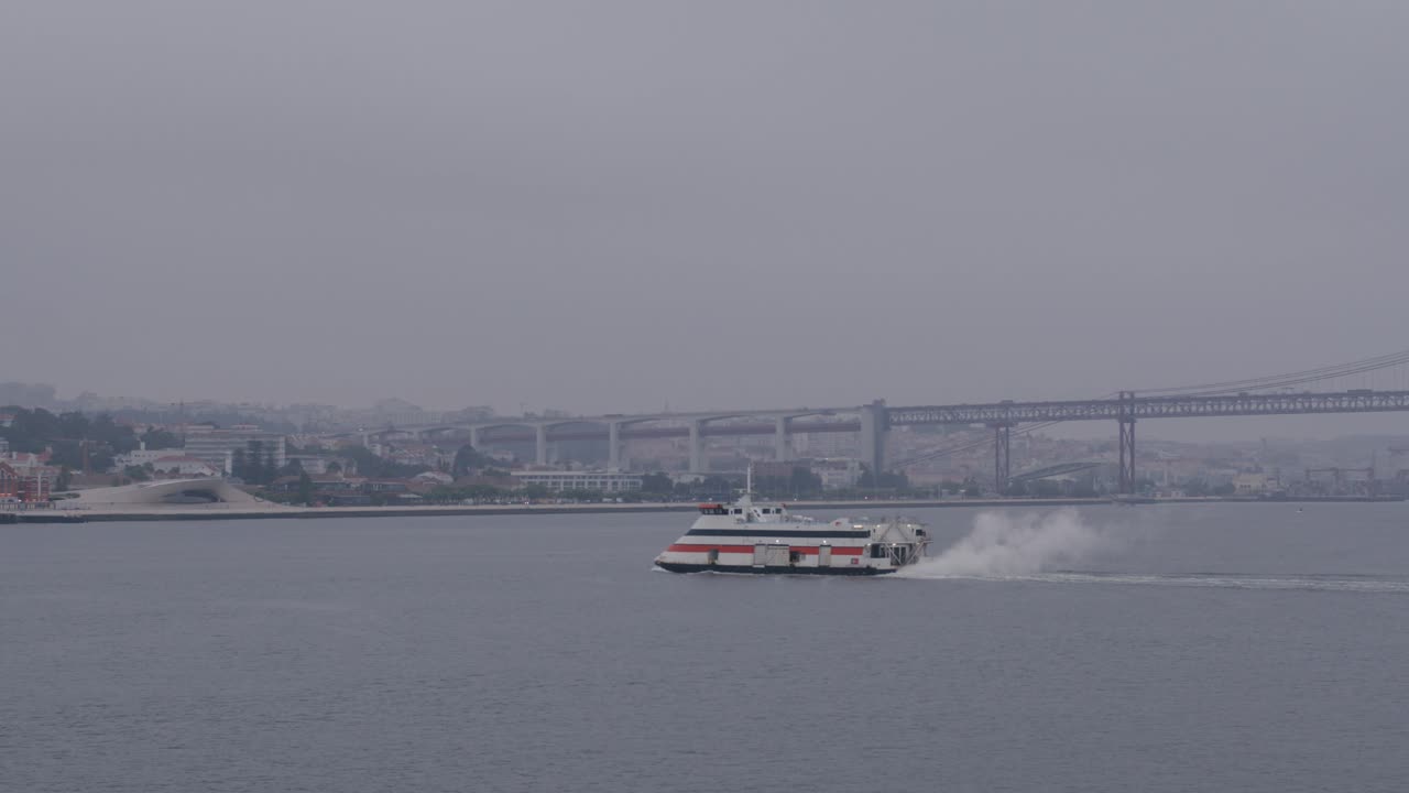 Ferry glides across the Tagus River in Lisbon on an overcast day, bridge in the background