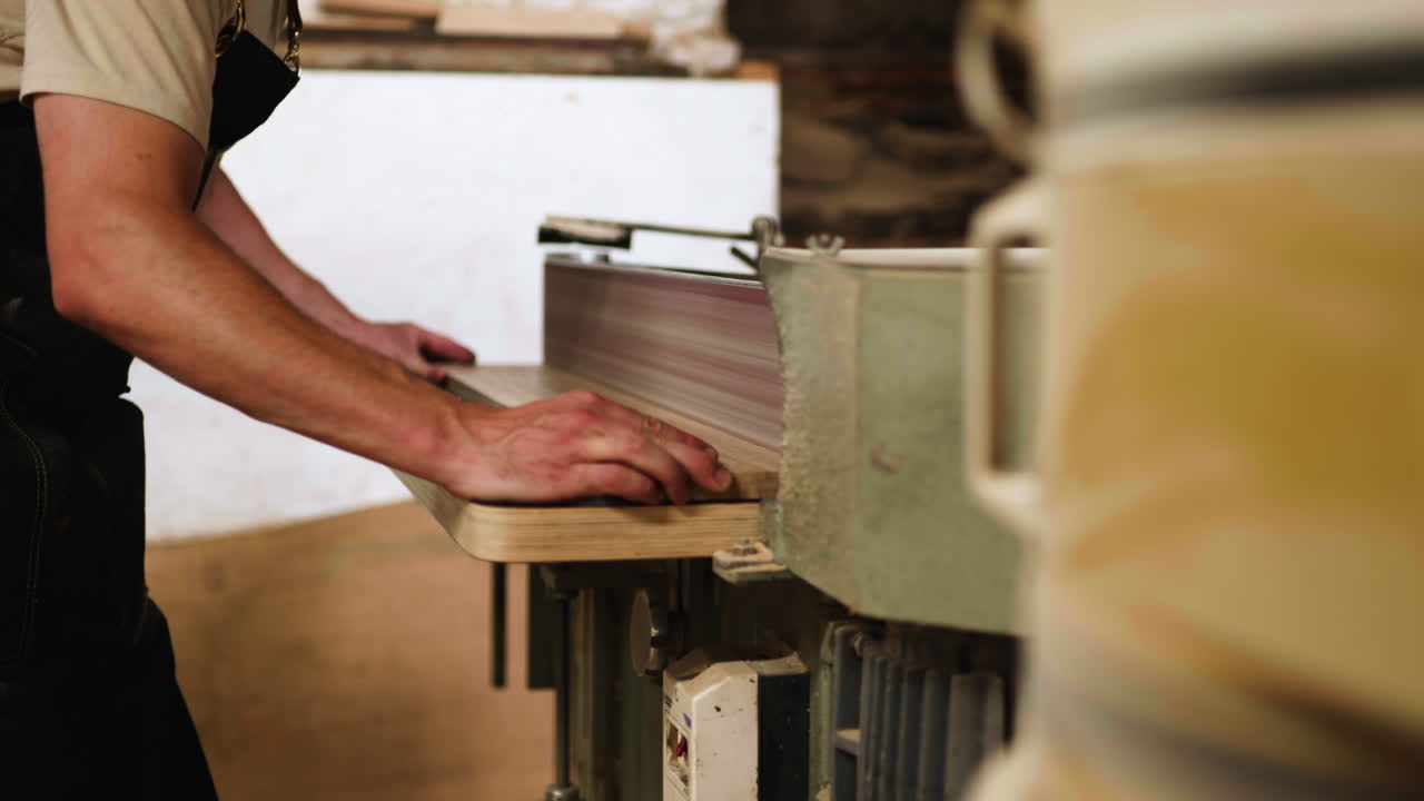 A carpenter focuses on shaping a wooden piece using a planer in a well-lit workshop. The meticulous work showcases craftsmanship and attention to detail in woodworking.