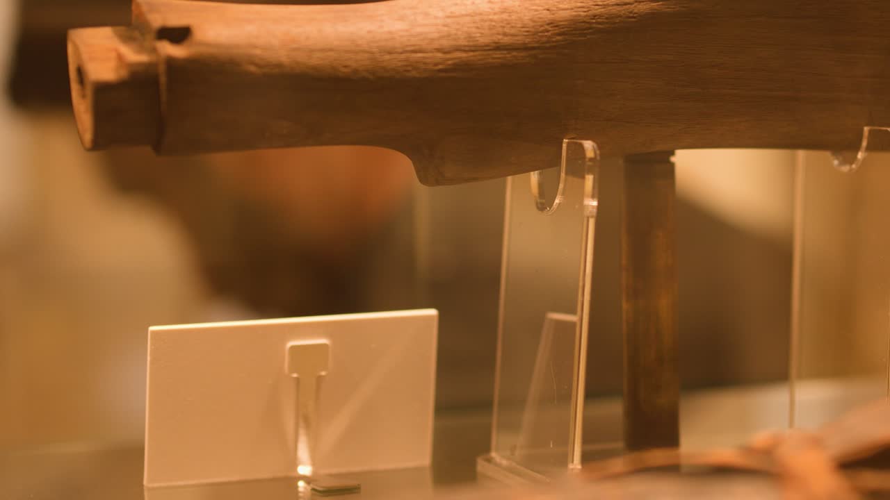 Warmly lit close-up of historical rifle stock, glass showcase, slow lateral camera movement, museum setting