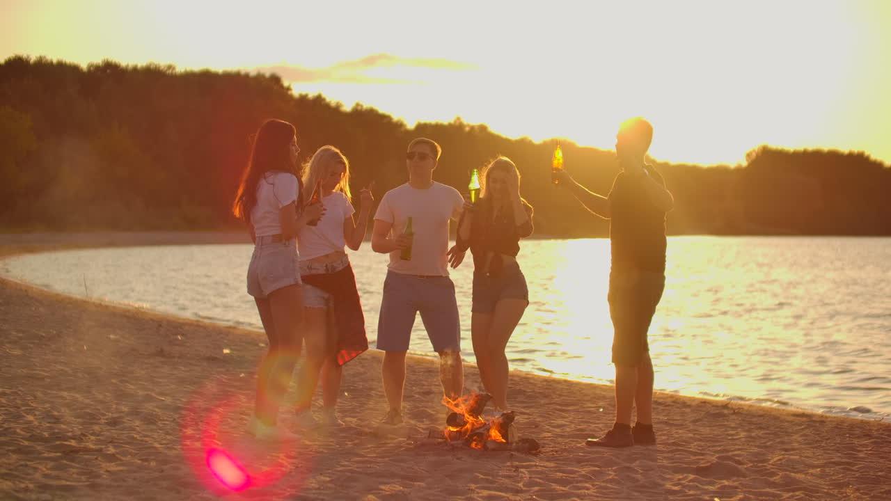 The students are dancing around bonfire on the beach with beer. They are talking to each other at sunset and enjoying the summer evening on the river coast.