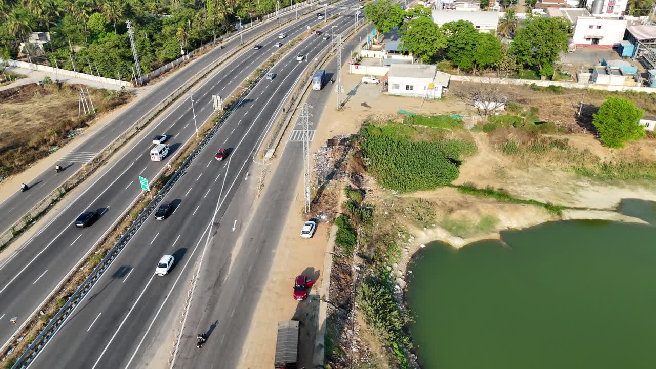 An aerial view showcases a multi-lane highway in India, Tamil Nadu, with vehicles, dry land, and a green-tinged water body. Urban structures and dense trees line the horizon. Service road