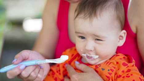 mother feeds her child sitting in her arms. baby eats white stuff and then closes eyes and frowns. close up.