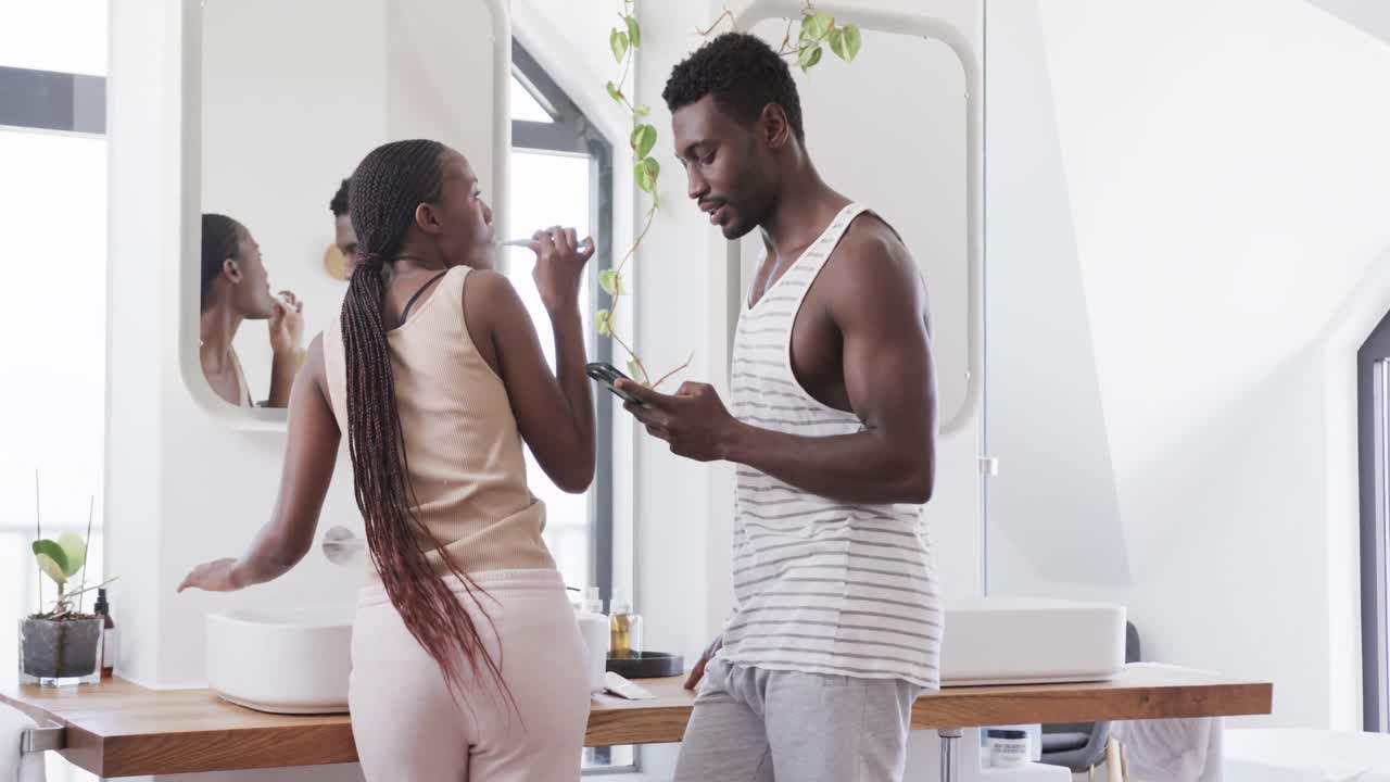 pareja afroamericana cepillándose los dientes, usando el teléfono inteligente y hablando en el baño, cámara lenta