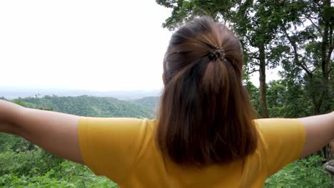 Camera-moves-back-revealing-the-head-of-a-woman-then-its-full-body-with-her-arms-stretched-out-as-she-is-wearing-a-yellow-shirt-welcoming-freshair-and-nature-itself-at-a-mountain-resort-in-Thailand
