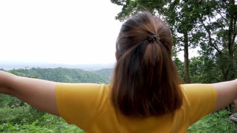 camera moves back revealing the head of a woman then its full body with her arms stretched out as she is wearing a yellow shirt welcoming freshair and nature itself at a mountain resort in thailand