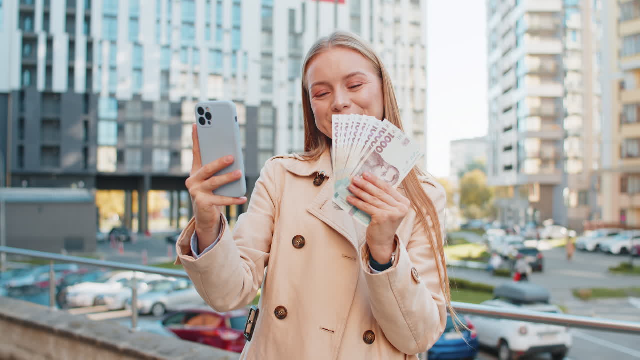 счастливая богатая зрелая бизнесменка использует смартфон, держит деньги, гривни, наличные на городской улице.