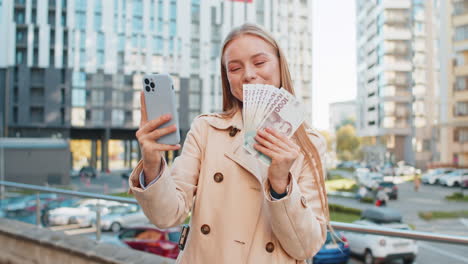 happy rich mature businesswoman lady using smartphone holding money hryvnias cash on city street