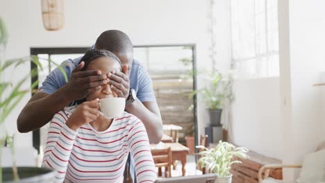 Happy-african-american-couple,-man-surprising-woman-at-table-in-sunny-coffee-shop,-slow-motion