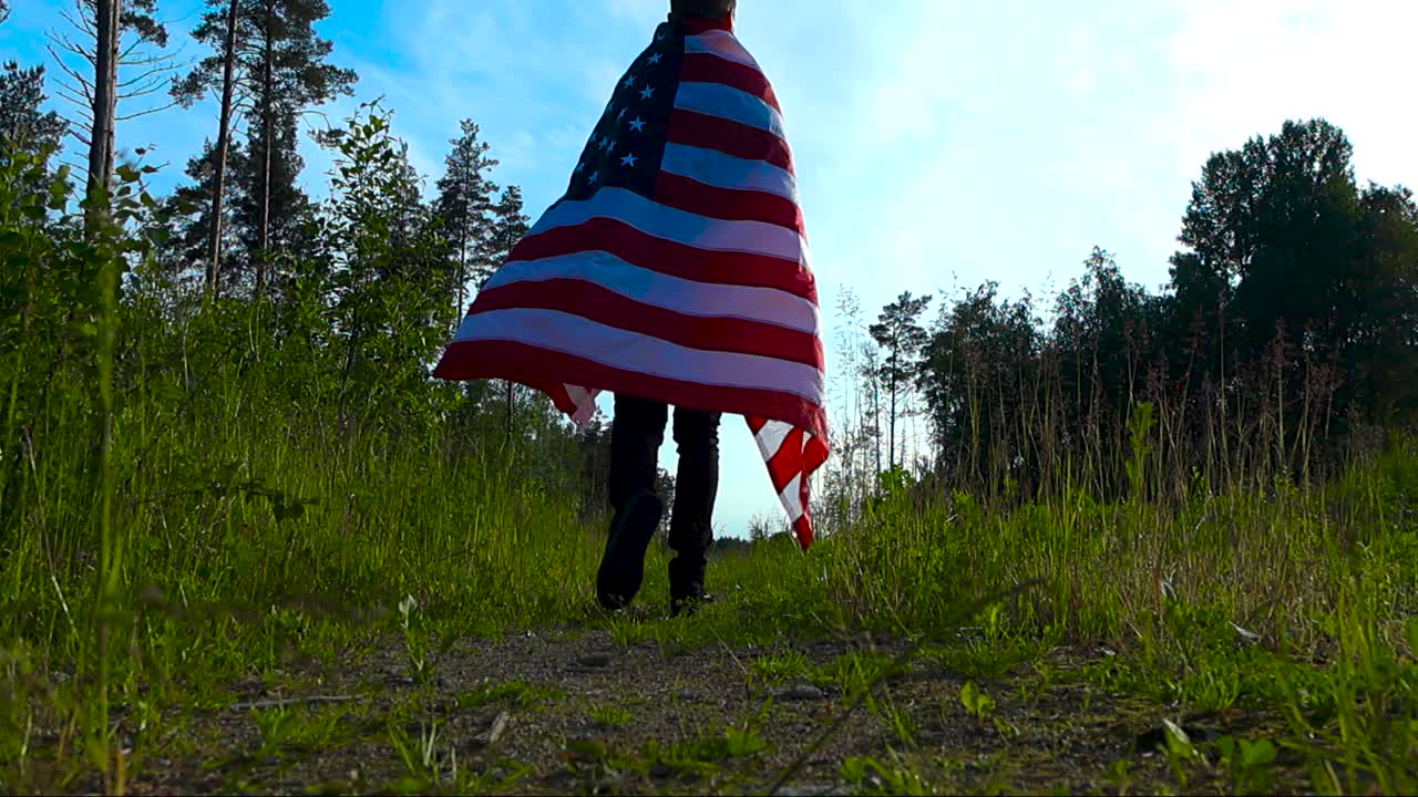 Gorgeous low angle video footage of a man stepping over a camera and walking into the distance on a rural countryside dirt road between tall grass while holding an American flag on his back, sunny day