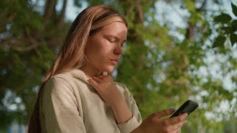 vista lateral de primer plano de una mujer con una mancha sutil en la cara, enfocada atentamente en su teléfono mientras sostiene su collar de capucha, la luz del sol se filtra a través de las hojas, con vegetación borrosa en el fondo