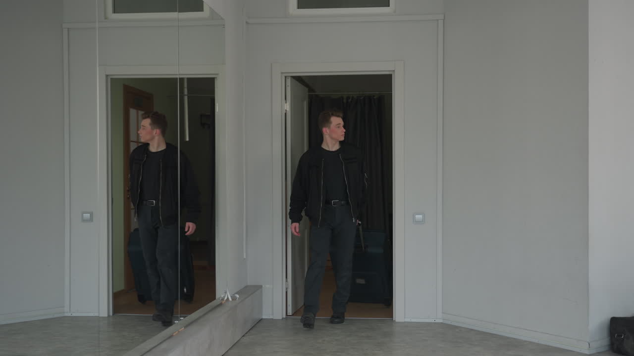 Young man stepping out of room holding suitcase, preparing to travel, visible through partially open door, with large vertical mirror reflecting room interior and background corridor