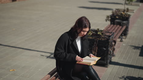 young woman in black coat sits on bench reading a book in sunlight, flipping to a new page, flower nearby sways gently with wind, and lamp post shadow stretches across paved ground in urban