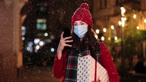 portrait of happy beautiful young woman typing on smartphone and holding a present on the street while it¬¥s snowing in christmas