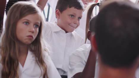 group of elementary pupils wearing uniform sitting on floor raise hands to answer question in class