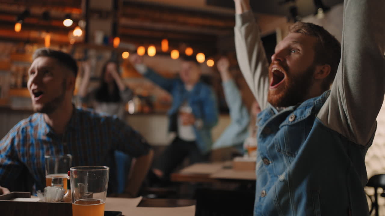 Close-up of a group of fans of men and women sitting together in a bar and watching a broadcast on TV enjoying a goal scored shouting and hugging. in football basketball.