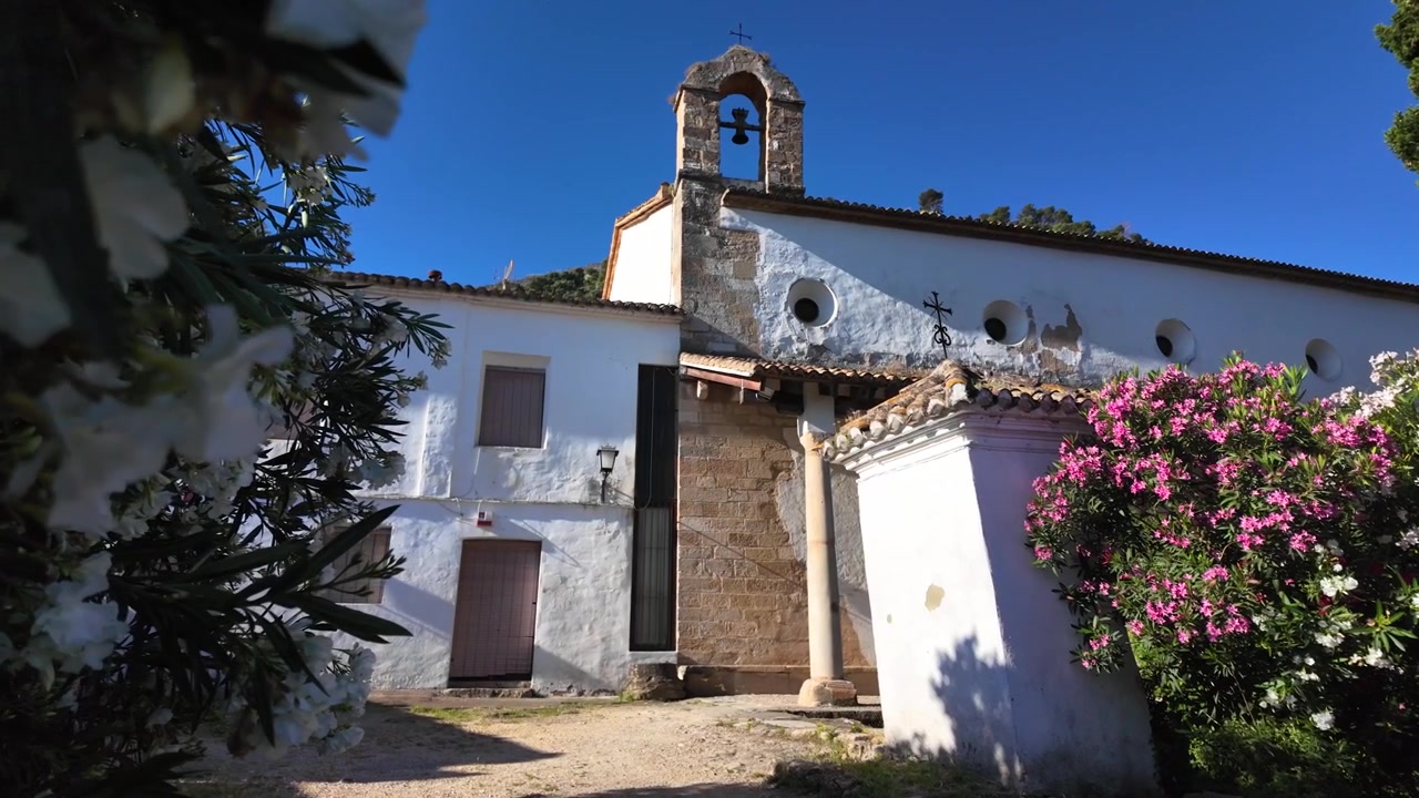 Revelación en cámara lenta de una iglesia tradicional encalada y edificios históricos cerca del Castillo de Xàtiva en Valencia, España, enmarcada por plantas con flores