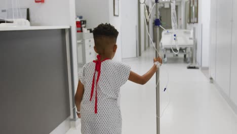 rear view of african american boy patient walking and holding drip stand in hospital corridor