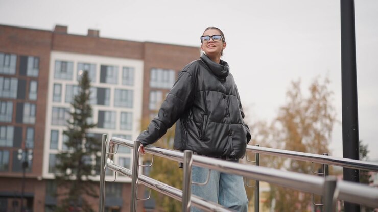 Close up of young woman wearing glasses and black puffer jacket standing on outdoor metal railing with urban buildings in background. Woman holds onto rail while looking around maintaining