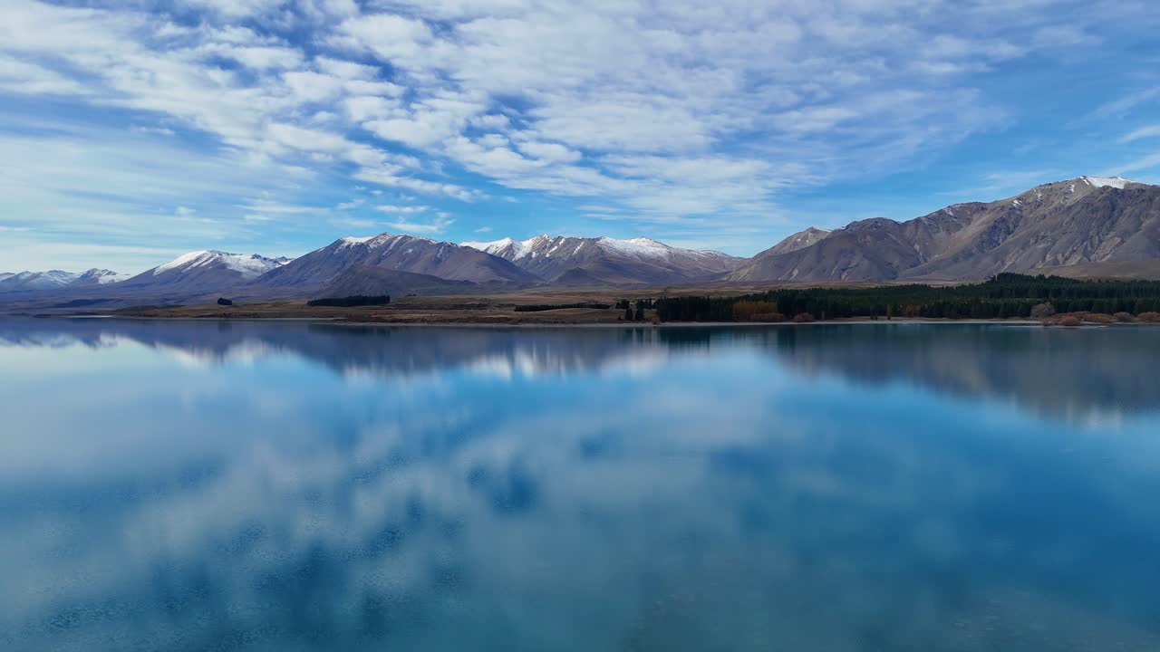 Drone footage captures Lake Tekapo's serene blue waters and surrounding mountains under a partly cloudy sky