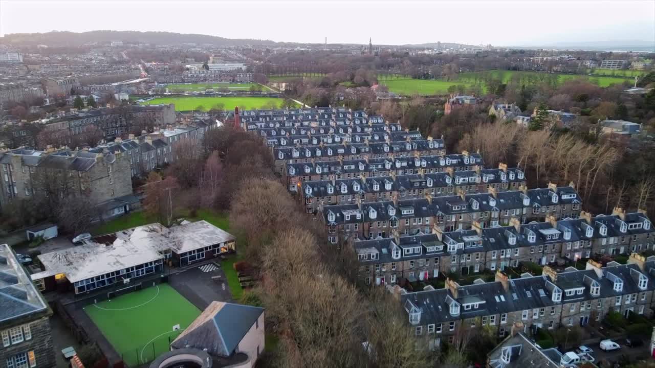 Footage of densely packed Edinburgh row houses, featuring a small sports facility and nearby green parks. Captures the classic stone architecture and the urban layout amidst surrounding greenery.