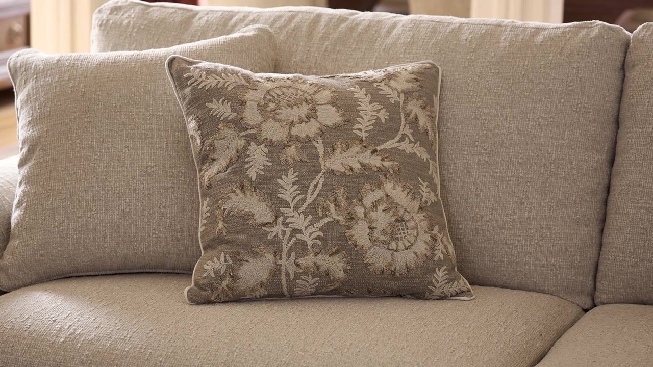 Långsam utzoomning av en ljusbeige blommig kudde på en grå soffa, belyst av varmt naturligt solljus i ett professionellt inrett rustikt vardagsrum.
