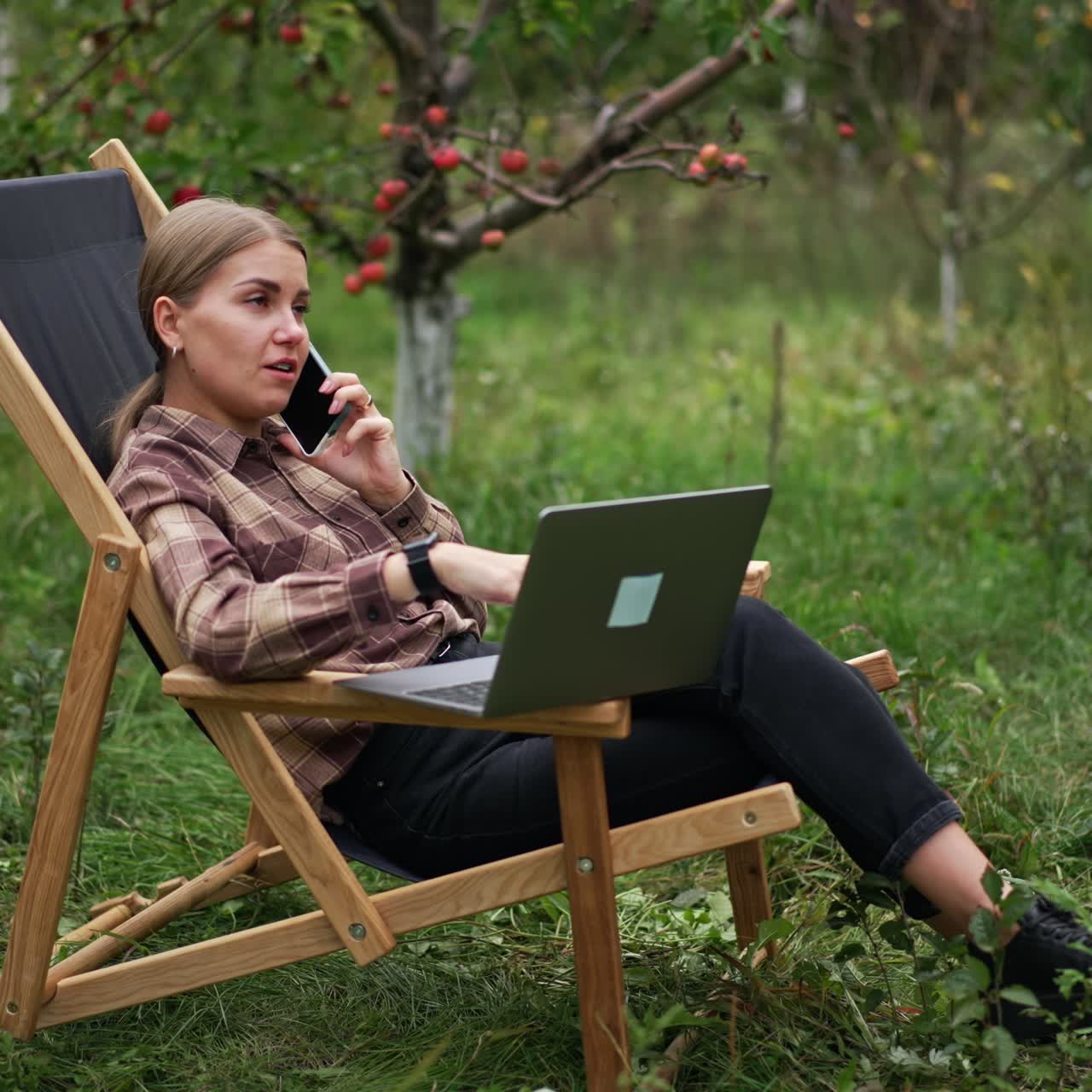 Freelance female specialist sitting in garden chair with laptop and phone. Lady is having conversation over the phone looking at computer screen