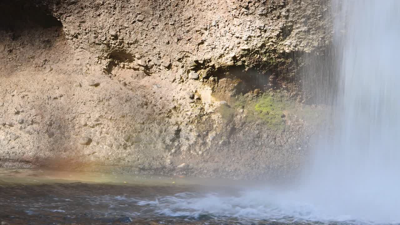 Sunlit waterfall cascades over mossy rocks, forming mist and a faint rainbow in natural setting