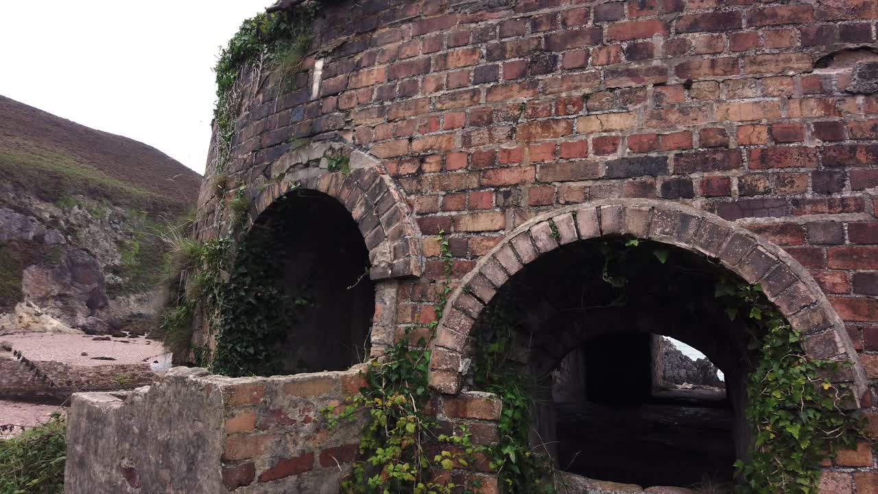 Ripresa all'indietro attraverso un arco delle rovine abbandonate e ricoperte di vegetazione di una fornace a cupola di mattoni gallese a Porth Wen