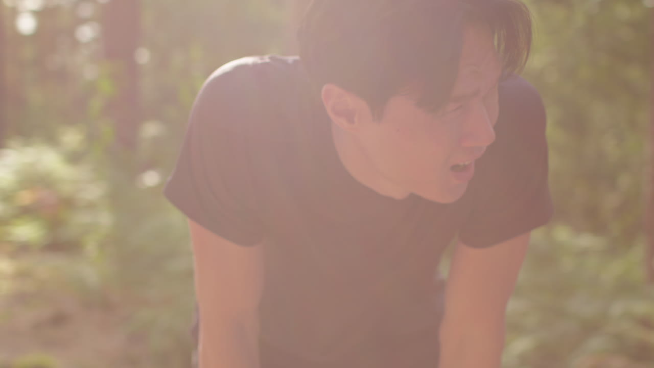 피곤한 중성인 남자가 야외에서 운동을 하고, 숲을 가로질러 달리고, 숨을 쉬기 위해 멈추고, 실시간으로 촬영합니다.