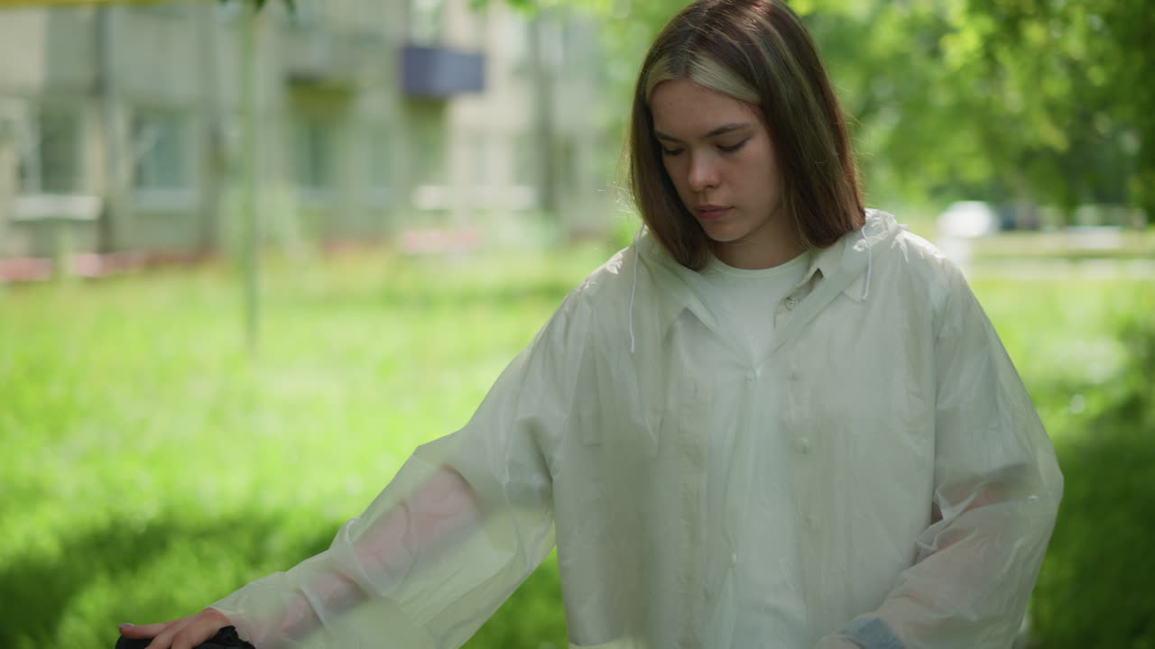 дама с светлыми волосами в полупрозрачном плаще останавливается на велосипеде на асфальтированной тропе, она выглядит капризно, окружена пышной зеленью с размытым видом зданий на заднем плане