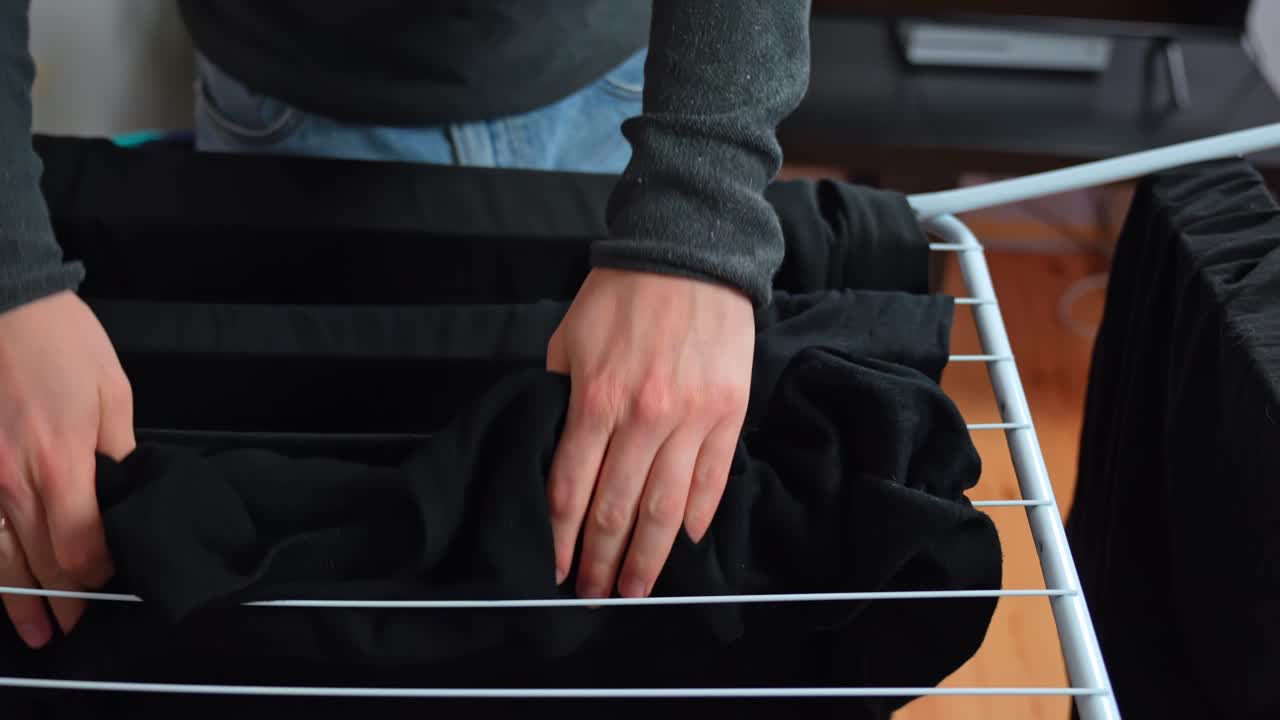 Close-up of a man hanging a black sweater on an indoor drying rack, arranging dark laundry neatly to air dry and save electricity in a small apartment