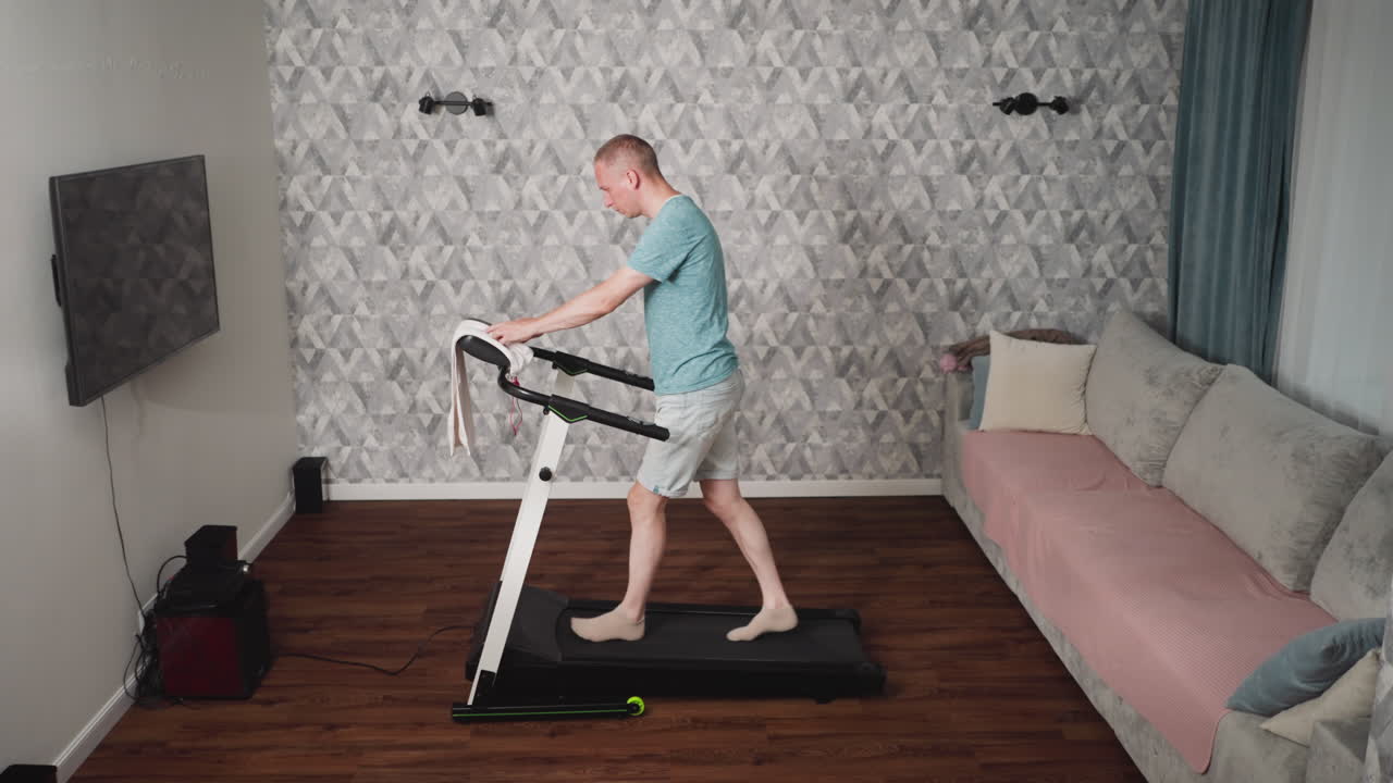 Young man in teal shirt and light shorts standing on treadmill adjusting towel draped on handle while preparing to increase speed during indoor workout in living room with patterned wall