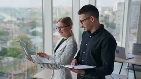 office colleagues talk looking at the screen of laptop in lady's hands. company employees discussing working moments using computer. blurred cityscape at backdrop.
