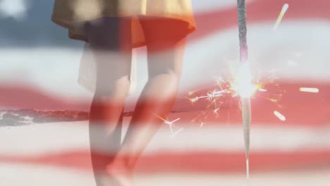 animation of american flag waving and sparkler over woman walking on beach