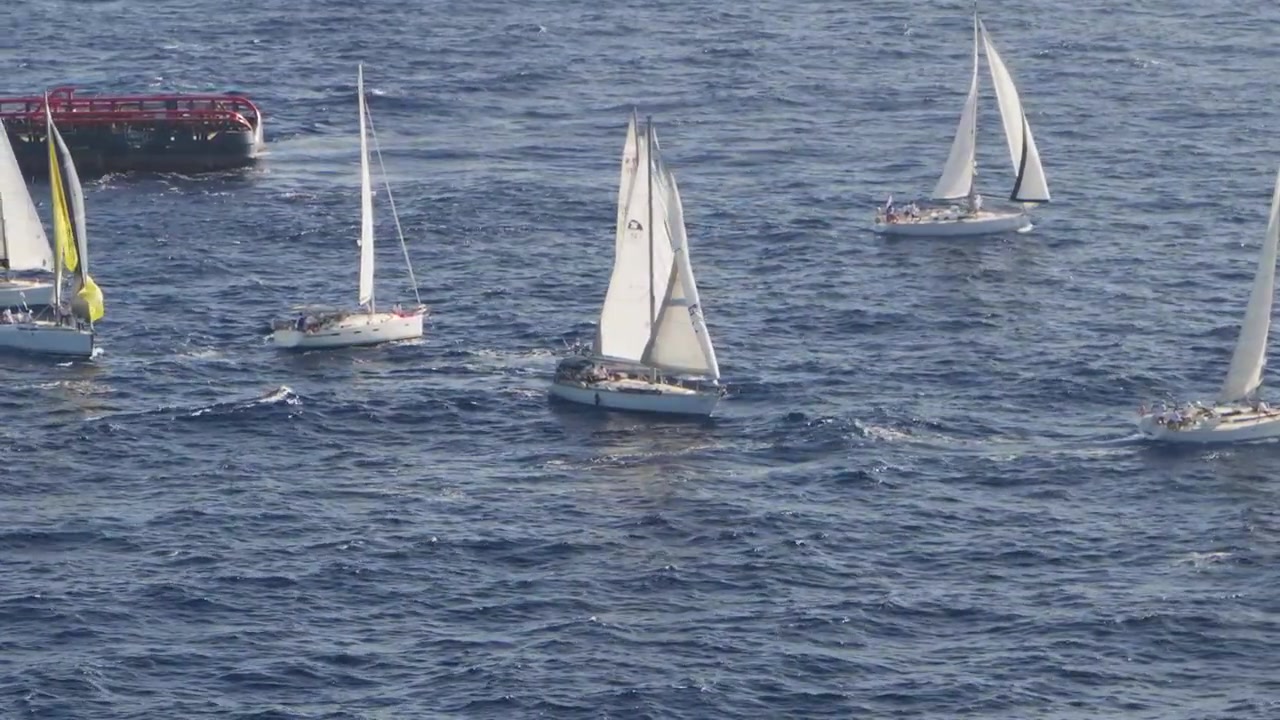 Vista dall'alto di più yacht che manovrano in stretta vicinanza durante una regata sul mare blu profondo con una barca di supporto nelle vicinanze.