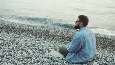 man sitting on a pebble beach contemplating the ocean