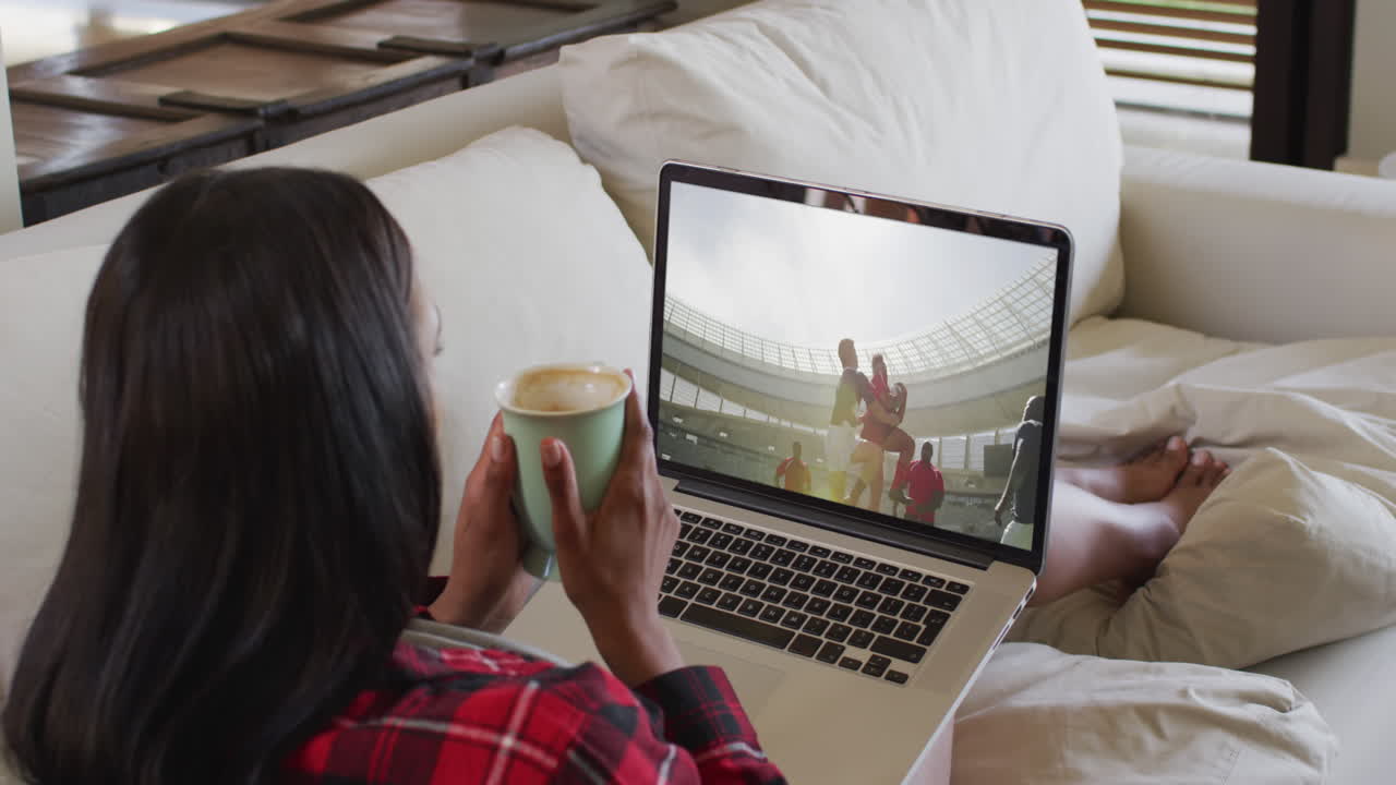 Composite of woman sitting at home holding coffee watching rugby match on laptop