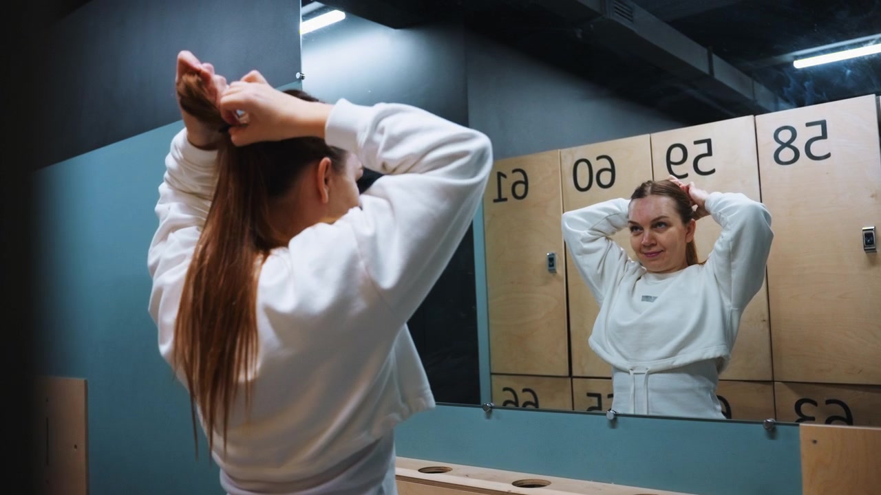 Mujer caucásica atándose una coleta frente al espejo con taquillas numeradas reflejadas, ajustándose la goma del pelo, luz cenital brillante, sonriendo a su reflejo, autoánimo y preparación tranquila antes del partido