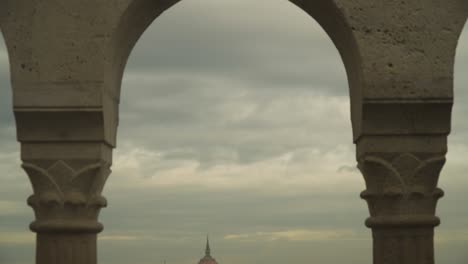 the hungarian parliament building and danube river captured between two concrete columns of a tall building structure in budapest hungary on a cloudy day - aerial shot