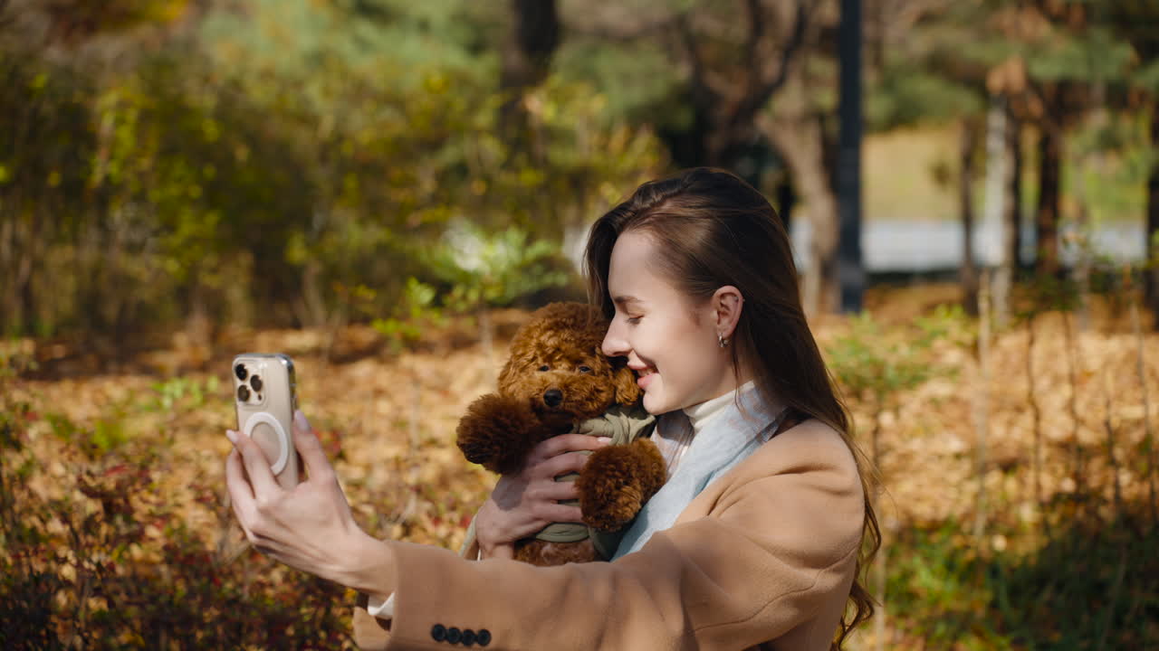 Cheerful Young Woman Taking Selfie Photo Holding Adorable Small Toy Poodle Puppy in Autumn City Park in Sunlight - orbit shot